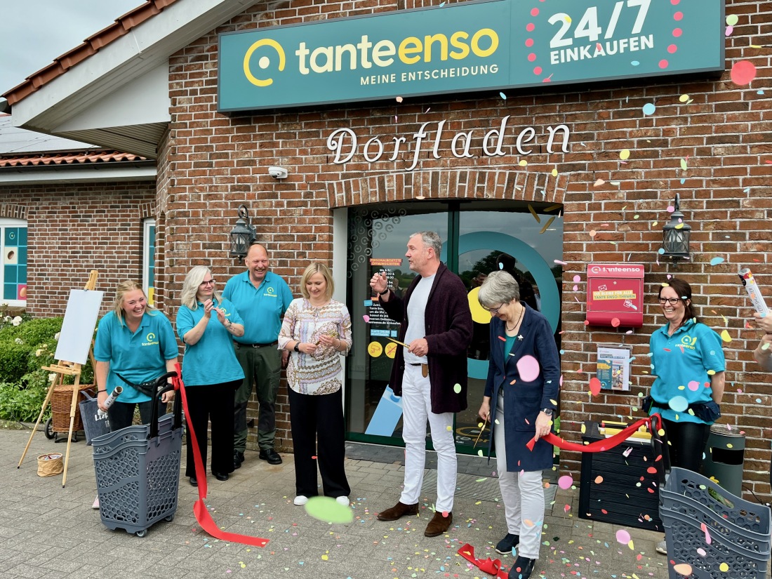 Sieben Personen stehen vor der Tante-Enso-Filiale in Holtsee zur Eröffnung. Es fliegt Konfetti, ein rotes Band wurde durchgeschnitten.