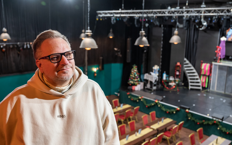 Ein Mann mit Brille in einem hellen Hoody steht oberhalb eines großen Bühnenraumes mit Tischreihen.