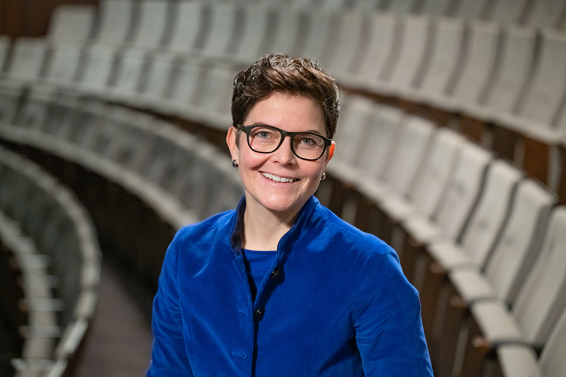 Eine Frau mit kurzen dunklen Haaren und Brille steht lächelnd zwischen den in Stufen angeordneten Stuhlreihen im Zuschauerraum eines Theatersaales.