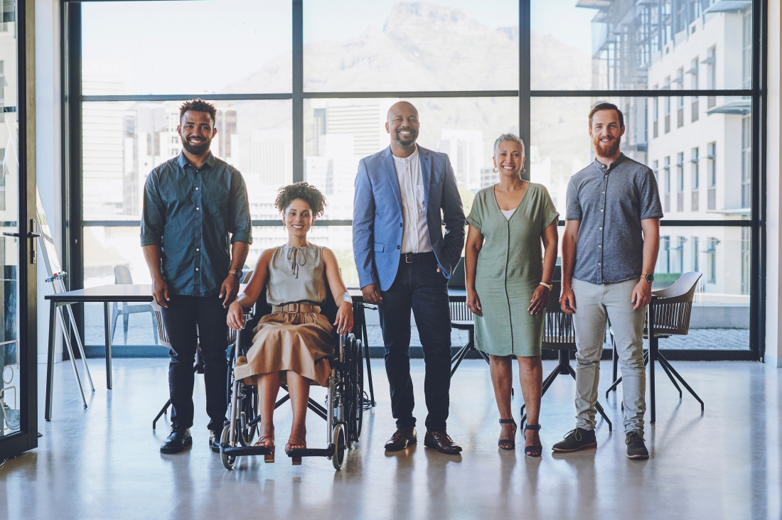 Eine diverse Menschengruppe (Hautfarbe, Geschlecht, Alter) in Business-Kleidung in einem moderen Bürogebäude vor einem Konferenztisch abgelichtet. Eine Frau sitzt im Rollstuhl. Eine diverse Menschengruppe (Hautfarbe, Geschlecht, Alter) in Business-Kleidung in einem moderen Bürogebäude vor einem Konferenztisch abgelichtet. Eine Frau sitzt im Rollstuhl.